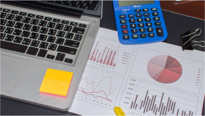 Desk setup for PMP certification preparation showing a laptop, financial charts, a calculator, and sticky