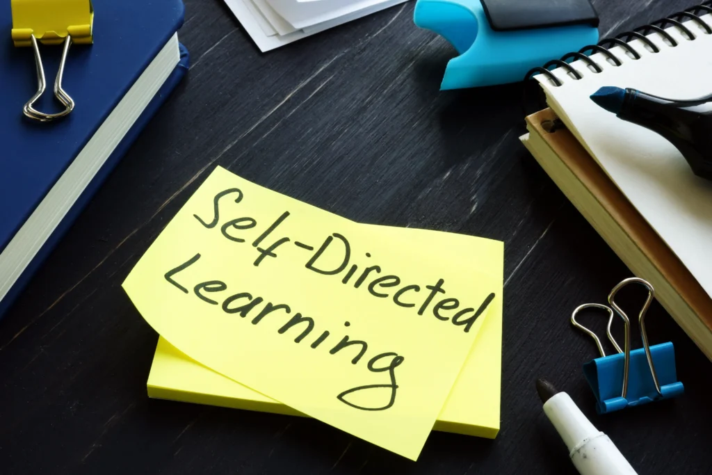  A sticky note labeled "Self-Directed Learning" surrounded by notebooks and stationery, representing the independence and risk of self-paced PMP courses.