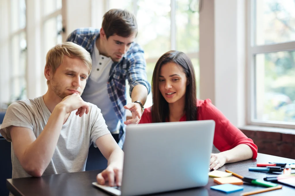 Three students collaborating over a laptop, researching PMP certification training options like Udemy and Education Edge.